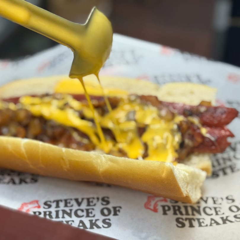 A hot dog with chili and cheese sauce being drizzled on top, on Steve's Prince of Steaks paper.