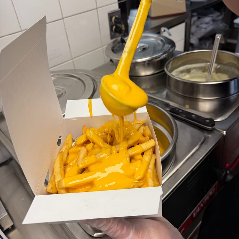 A yellow ladle pours melted cheese over a box of golden french fries in a restaurant kitchen.