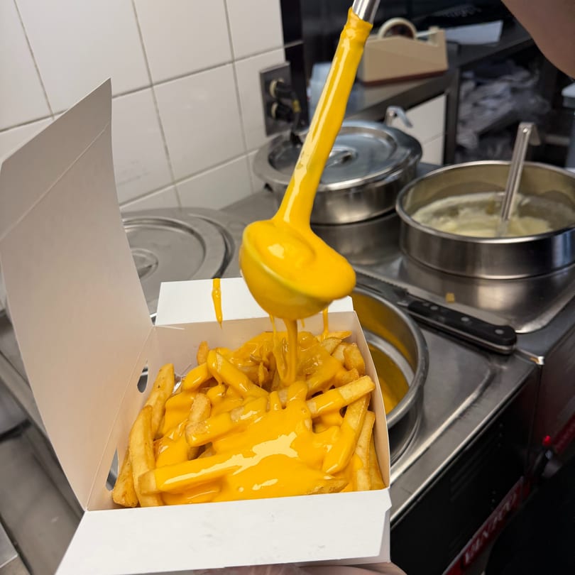 A ladle pours bright orange cheese sauce over a box of golden french fries in a commercial kitchen.
